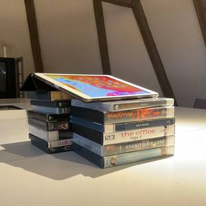 Ein Tablet auf einem Stapel DVDs, im Hintergrund ein Buch und ein Fernseher