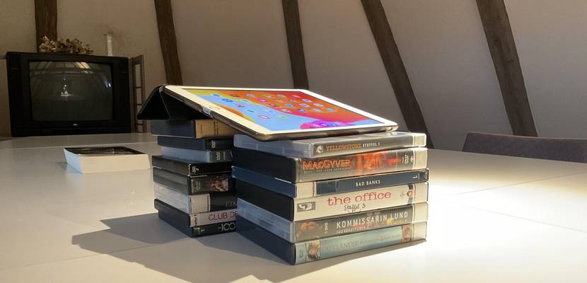 Ein Tablet auf einem Stapel DVDs, im Hintergrund ein Buch und ein Fernseher